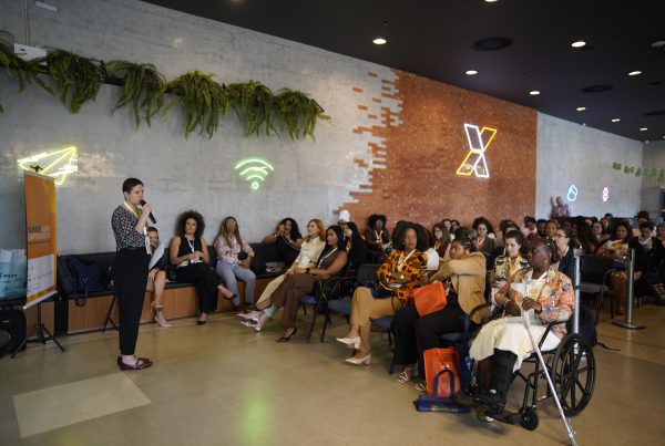 Uma pessoa está em pé, falando ao microfone para uma plateia durante um evento de empreendedorismo. O público, formado por dezenas de participantes, está sentado em cadeiras e acompanha atentamente a apresentação. Em primeiro plano, uma participante utiliza cadeira de rodas e muletas. O ambiente é moderno, com parede de concreto e tijolos aparentes, plantas suspensas e letreiros de neon na parede. À esquerda, há um banner do Summit Aliança Empreendedora. Os participantes usam crachás, indicando que se trata de uma atividade de conferência ou encontro profissional.