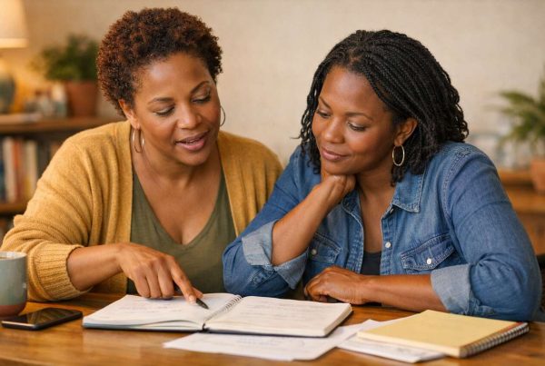 Duas mulheres estudam e colaboram em mesa de madeira, revisando anotações, café ao lado, ambiente acolhedor, simbolizando aprendizado e humanismo.