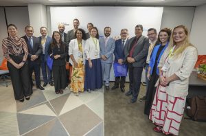 Foto de grupo com 14 participantes de um encontro institucional. As pessoas estão lado a lado, sorrindo para a câmera, usando crachás de identificação e trajes sociais ou esporte fino. Ao fundo, há um painel com as marcas da Fundação Arymax, Fundação Volkswagen e do Summit Aliança Empreendedora. O ambiente é uma sala de eventos, com iluminação interna e mesas laterais com materiais do encontro.